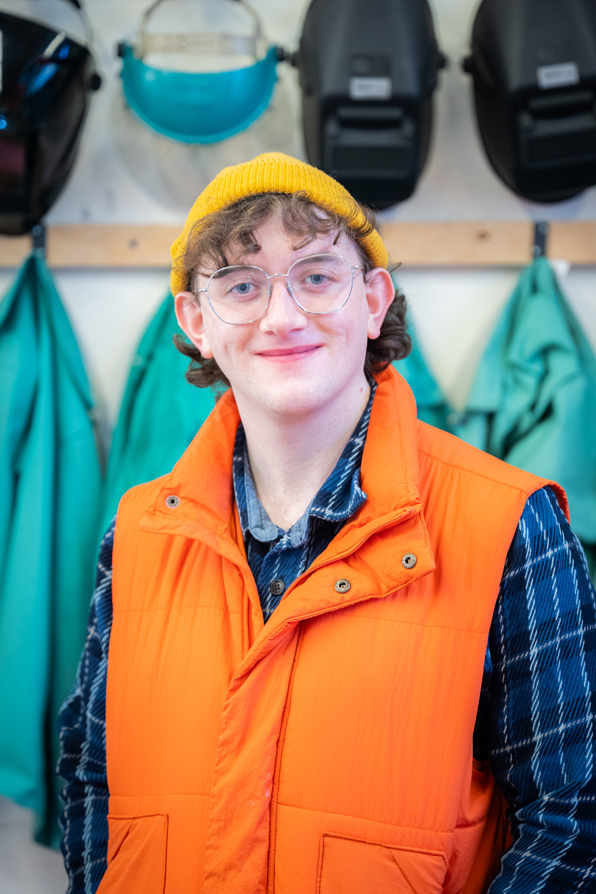 James is smiling looking at the camera. James is wearing a yellow beanie, glasses and a blue plaid shirt under an orange vest.