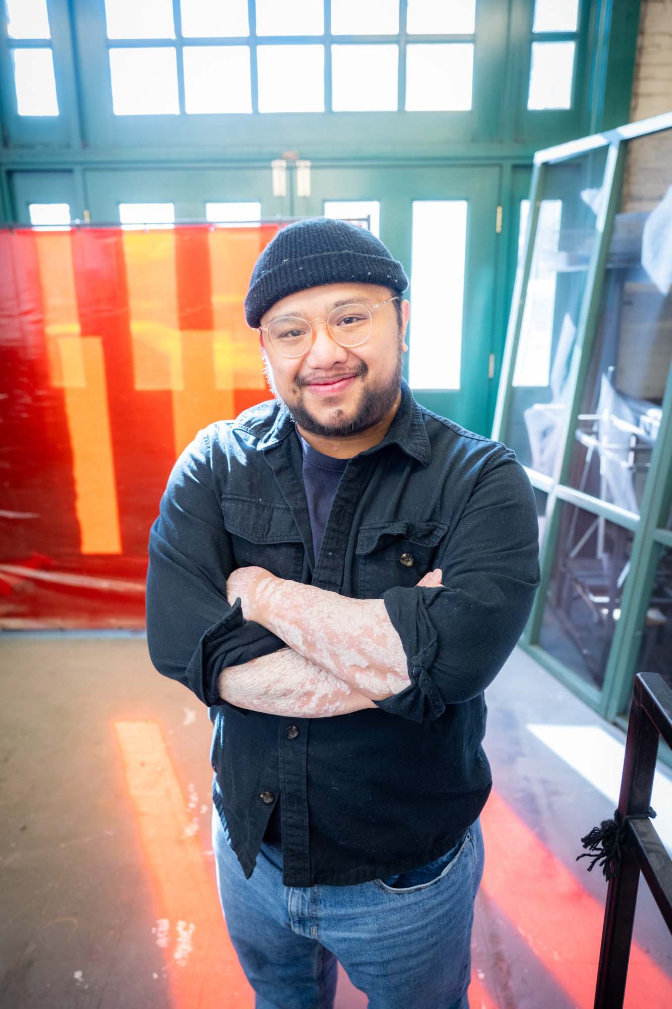 Reed is wearing a black beanie, glasses and a black shirt. Reed is smiling looking at the camera with their arms crossed.