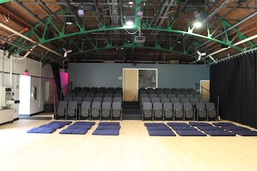 6 Rows of cushioned theatre seats with an aisle down the middle. In front are 3 rows of large pillow cushions placed on the floor.