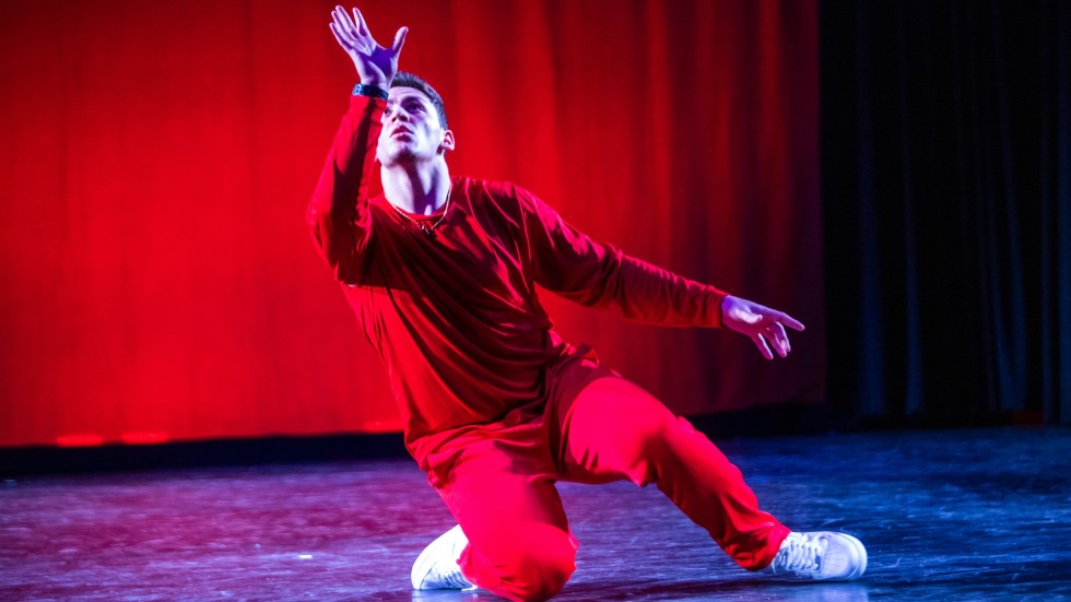 A man wearing all red dancing in the FlexN style. One knee is on the floor and one hand is up in the air. 