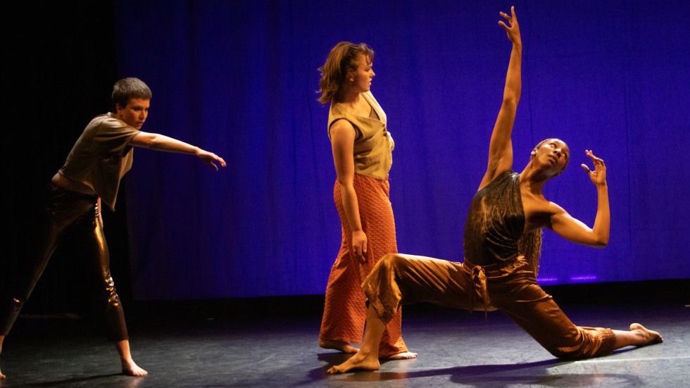 Three dancers in various poses. The person to the left is standing with their left arm crossed in front of them. In the center someone is standing slightly tilted to the side. To the right is someone with one knee bent behind them. 