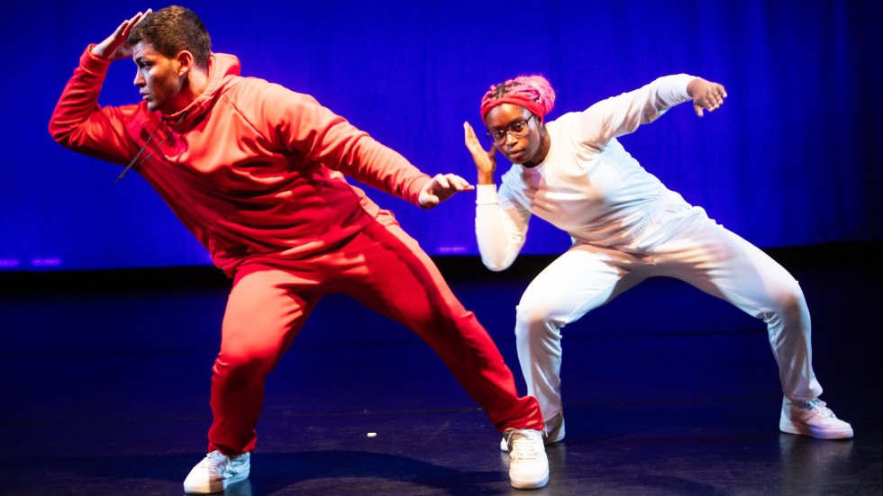 Two Dancers, both have their left hand on their head and their right hand out towards the right. They are both slightly bent at the knees. 