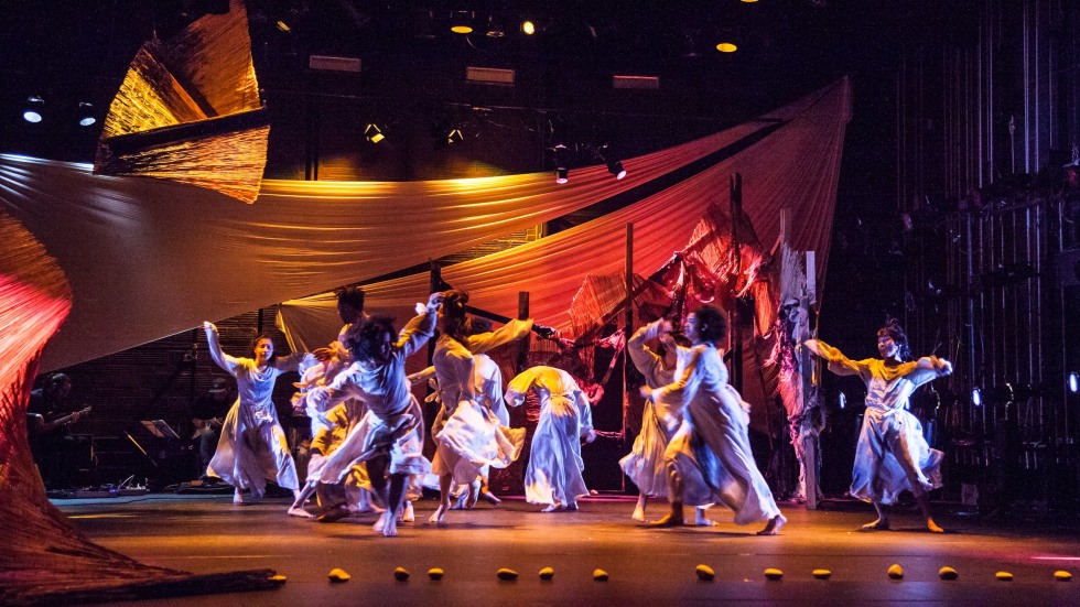 A large group of dancers on stage with fabric hung above them. 