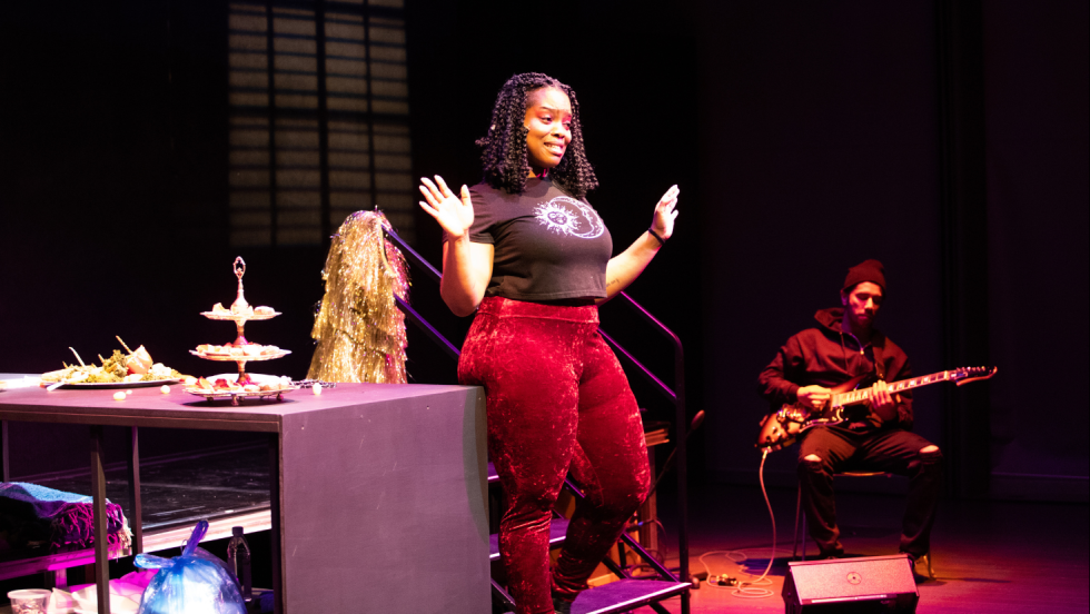 In the foreground is a woman wearing red pants and a graphic t-shirt. In the background is a person sitting playing the guitar. 