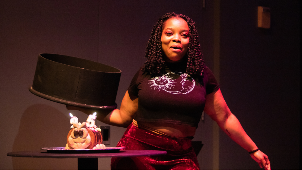 A woman lifting a lid to reveal a birthday cake with "2" and "5" candles lit in it.