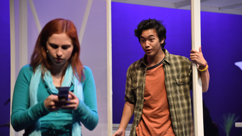 A young woman busy with her phone, a young man in the background, behind her facing her. 