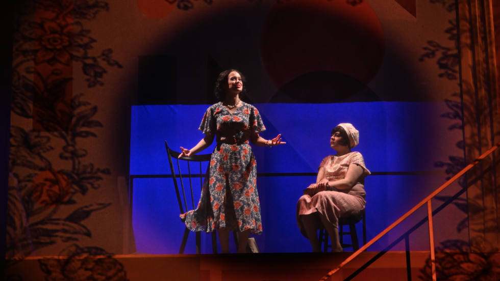 Two women in spotlight, one is standing and one is sitting down on a chair. 