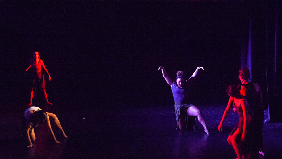 A young woman in center stage kneeling on the floor with arms stretched up over her head, 4 other dancers in the background surrounding her.