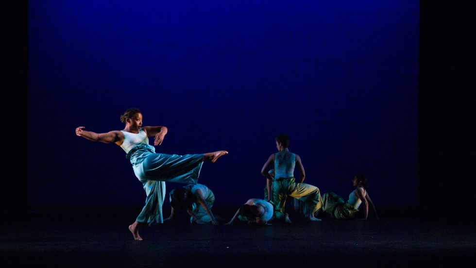 A dancer with her leg upwards and five dancers dancing in the background 