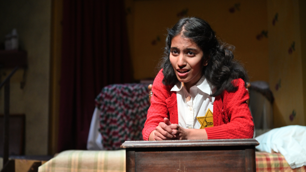 Young woman wearing a red cardigan and a yellow Star of David pinned to her chest. She is sitting at a desk looking outward.