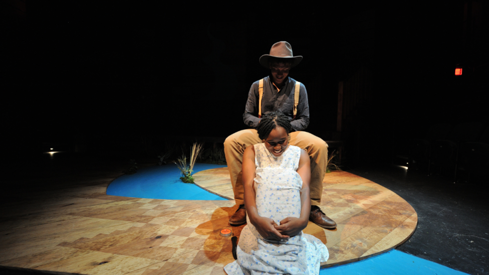 A young woman sitting on the floor in between the legs of a man sitting on a chair. He is braiding her hair. 