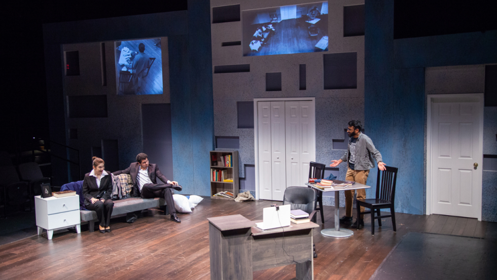A man standing at a small dining table talking to a man and woman sitting on a couch across from him. 