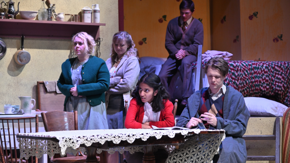3 women and 1 man gathered at a dining table. A man sitting on a bed in the background.
