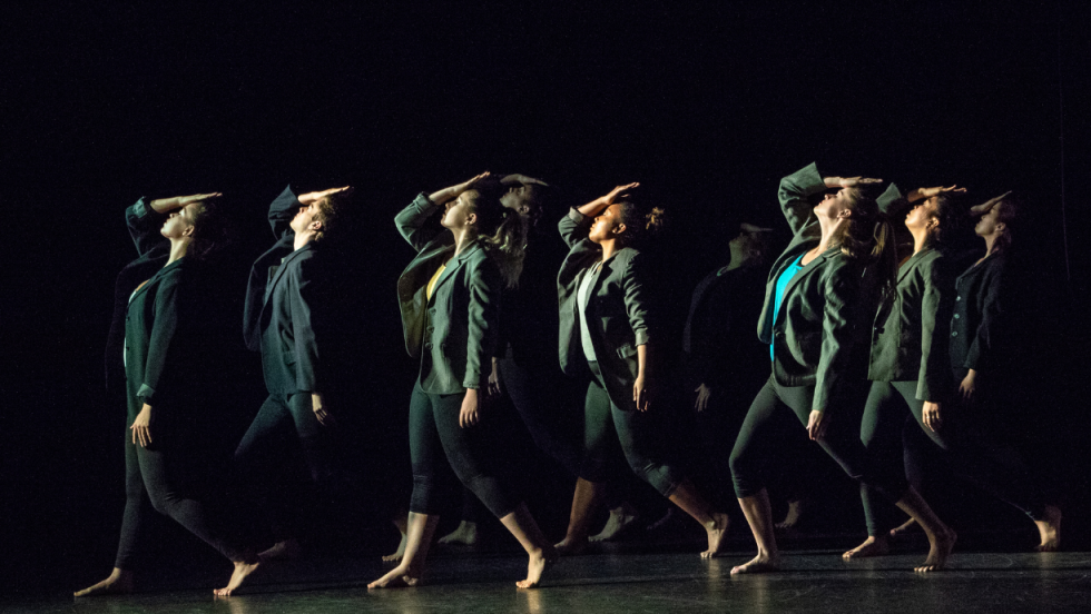 Sideview of a group of dancers dancing with their heads tilted back and their hands on their heads. 