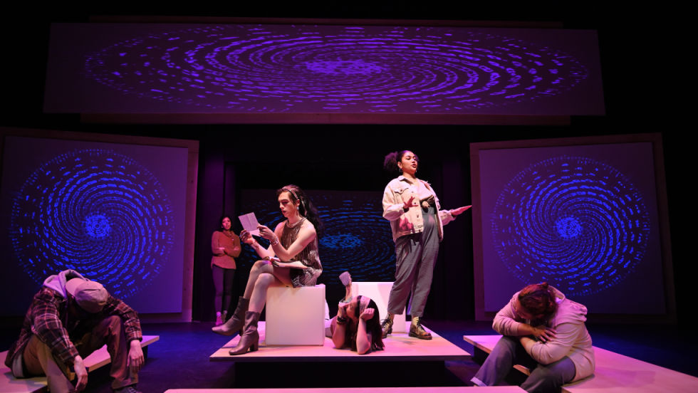 3 women in center stage, 1 sitting on a block, on laying the floor and the third woman standing. 3 people on the outer parts of the stage 