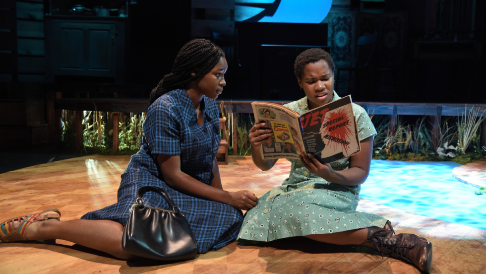 Two young women sitting on the floor. One is holding a magazine and reading it, the second is looking at her. 