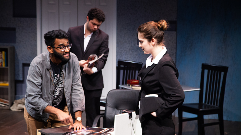A man at a desk talking to a woman, a young man flipping through a magazine in the background. 