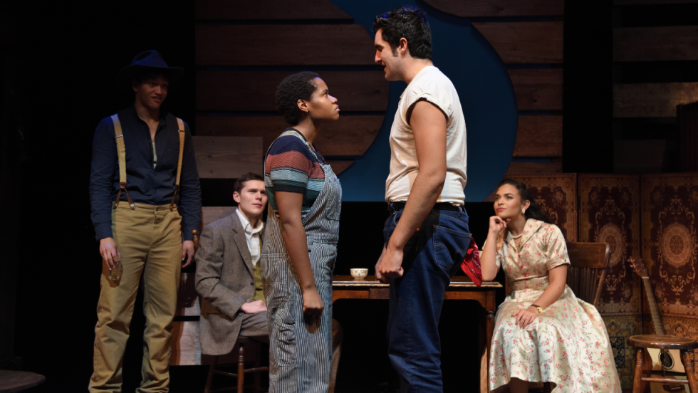 A man and a woman standing and looking at each other sternly. 3 people looking on in the background. 