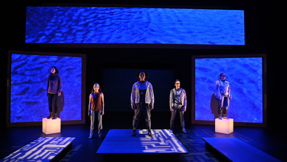 5 people standing facing the audience on a dimly lit stage. 
