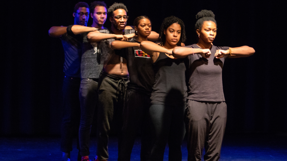 3 women and 3 men in a line facing the audience with their arms raised horizontally across their chest and hands in fists. 