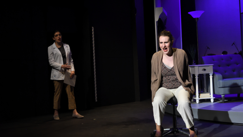 A woman sitting on a chair, a woman in the background facing her.