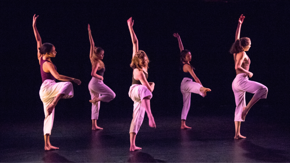 5 female dancers in a dance with one arm raised above their heads, standing on one leg. 