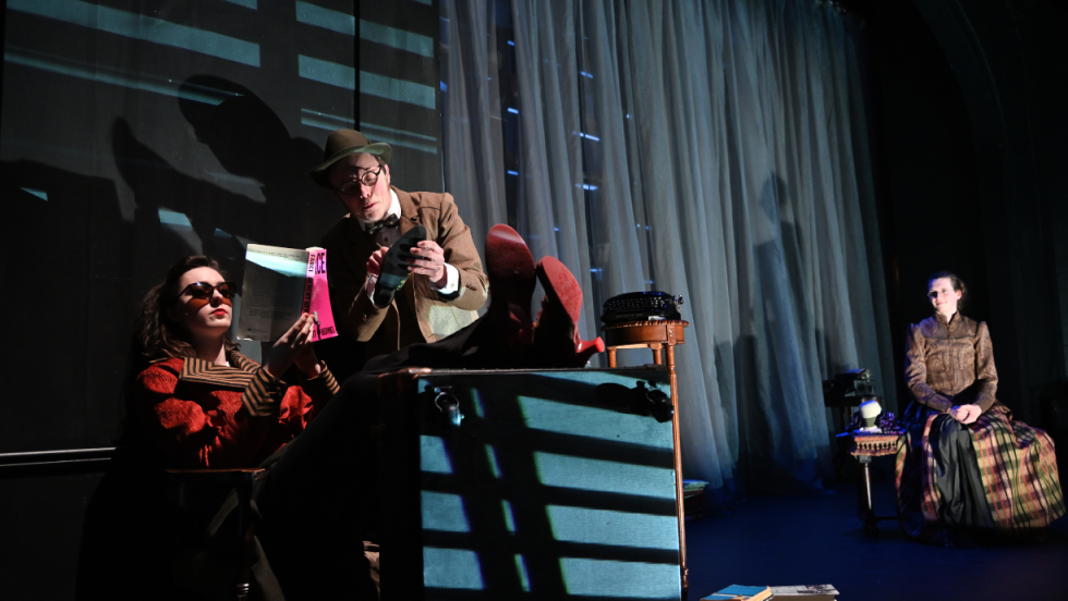 A young woman reading a book with her feet perched on some furniture, a man trying to talk to her, and a woman in the background watching them. 