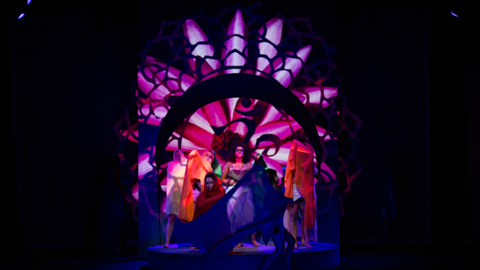 A group of dancers draped in sheer colorful fabric in center stage. 