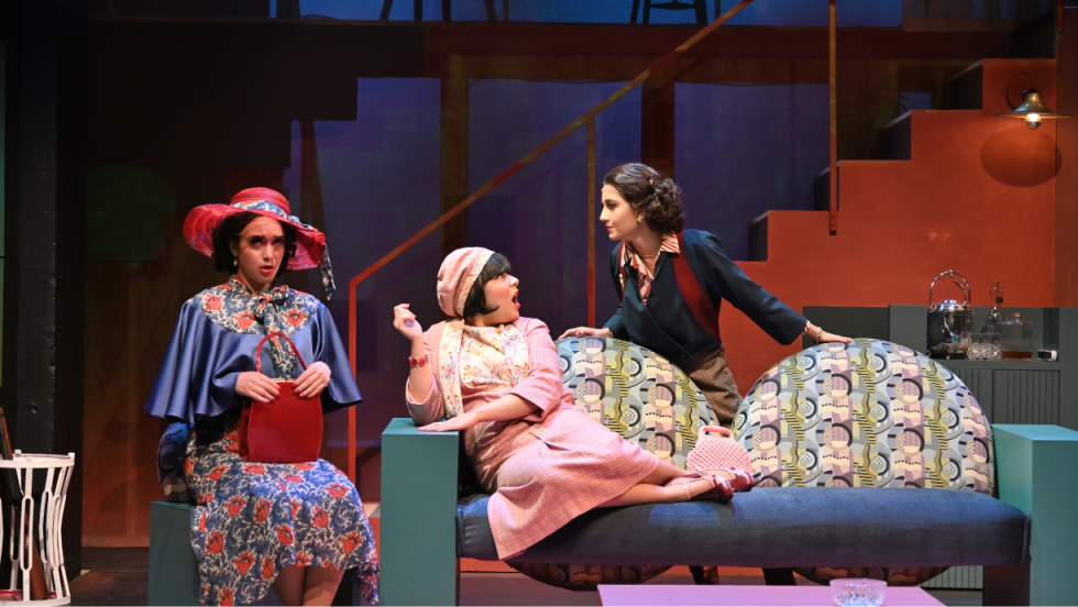 3 women together, one is sitting on a futon, one standing behind the futon, the third woman sitting in a chair next to the futon. 