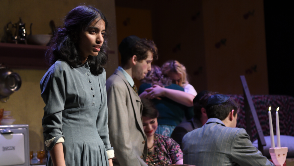 A young woman looking toward the audience, two women embracing in the background, and 4 people in the background looking sullen. 