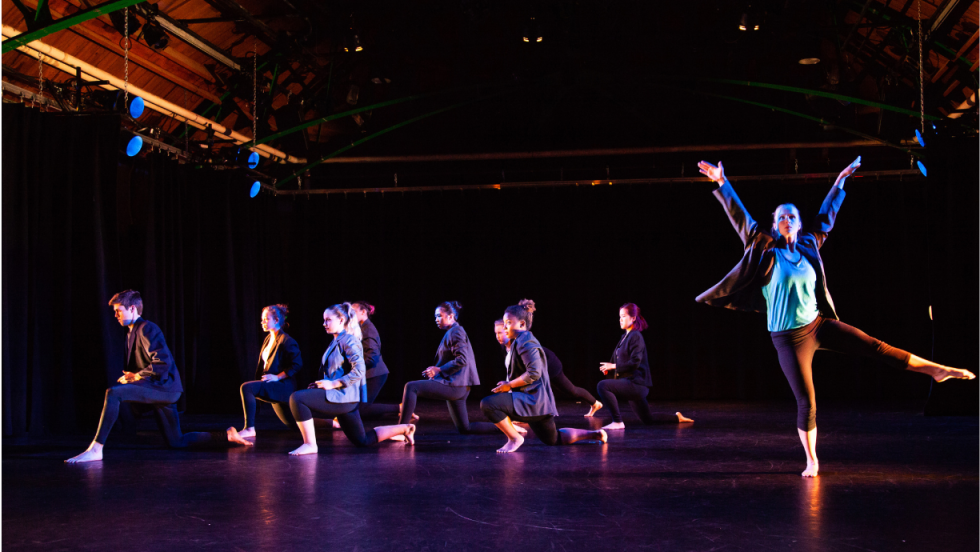 A female dancer dancing, ad balancing on one leg, and other dancers kneeling in the background