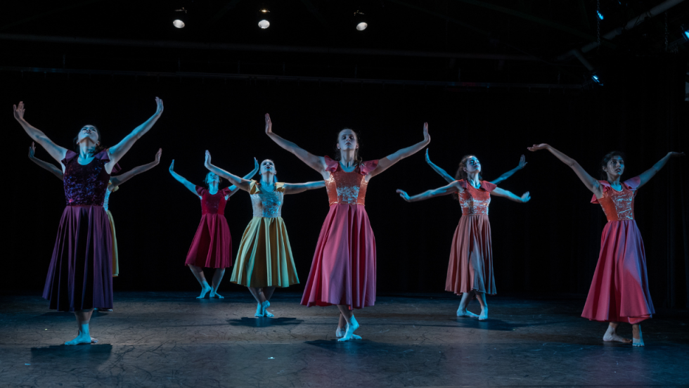 A group of female dancers in colorful dresses with their arms raised high above their heads and their feet crossed. 