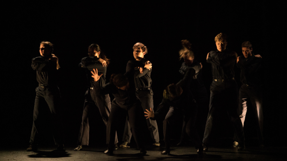 A group of dancers in black, dancing vigorously in dim lighting 