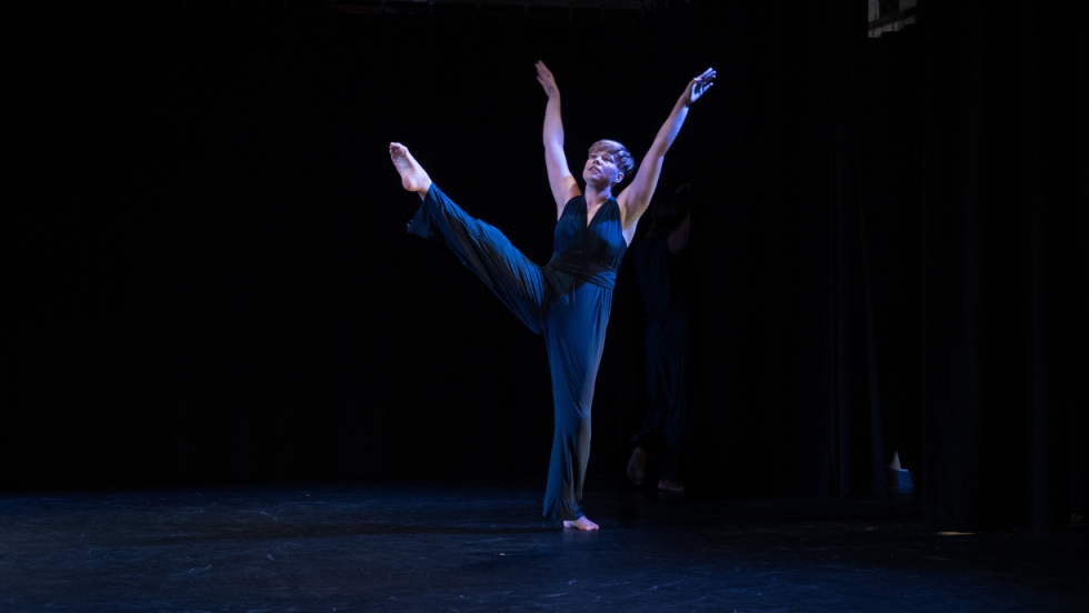 A solo dancer in dim lighting dancing with one of her legs up and both arms outstretched above her head.